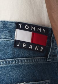 Jeans de denim con un lavado azul, presentando una etiqueta de tela rectangular en azul marino con rectángulos blancos, rojos y azul marino, y "TOMMY JEANS" en texto blanco.