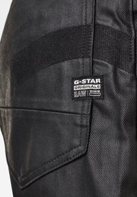 Černé džíny s texturovaným povrchem, šikmou zadní kapsou a štítkem G-Star Originals s nápisem "RAW DENIM" v bílé barvě.