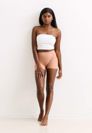 Frau mit dunklem Haar, die ein weißes trägerloses Top und beige Shorts mit hohem Bund trägt, barfuß vor einem schlichten hellen Hintergrund steht.