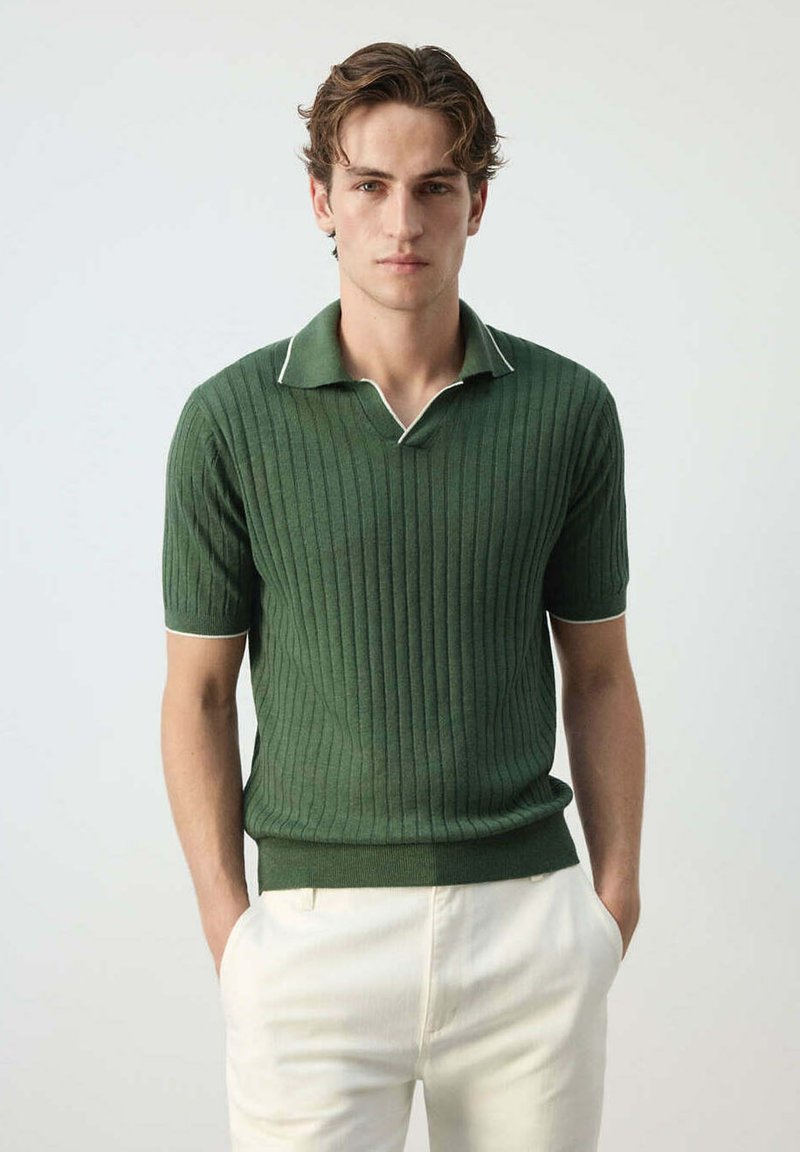 Jeune homme aux cheveux bruns portant un polo vert côtelé à manches courtes et un pantalon blanc, debout les mains dans les poches sur un fond clair.