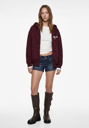 Giovane donna che indossa una felpa con cappuccio bordeaux con zip, una maglietta corta bianca, pantaloncini in denim scuro e stivali alti in pelle marrone, in piedi su sfondo bianco.