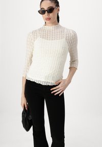 Top din dantelă de culoare crem, cu mâneci trei sferturi, guler bufant și tiv ondulat, asortat cu pantaloni negri și un clutch negru.