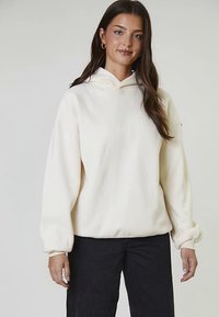 Sudadera con capucha de color crema en tela suave con puños y dobladillo acanalados, combinada con jeans negros. Diseño sencillo y relajado sin estampados.