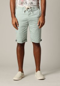 Man in opgerolde lichtgroene shorts met trekkoord en witte instappers, staand tegen een effen achtergrond.