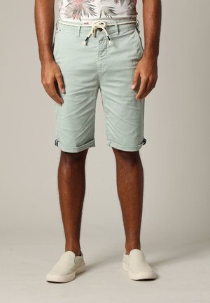 Homme portant un short vert clair roulé avec un cordon de serrage et des chaussures blanches sans lacets, se tenant devant un fond uni.