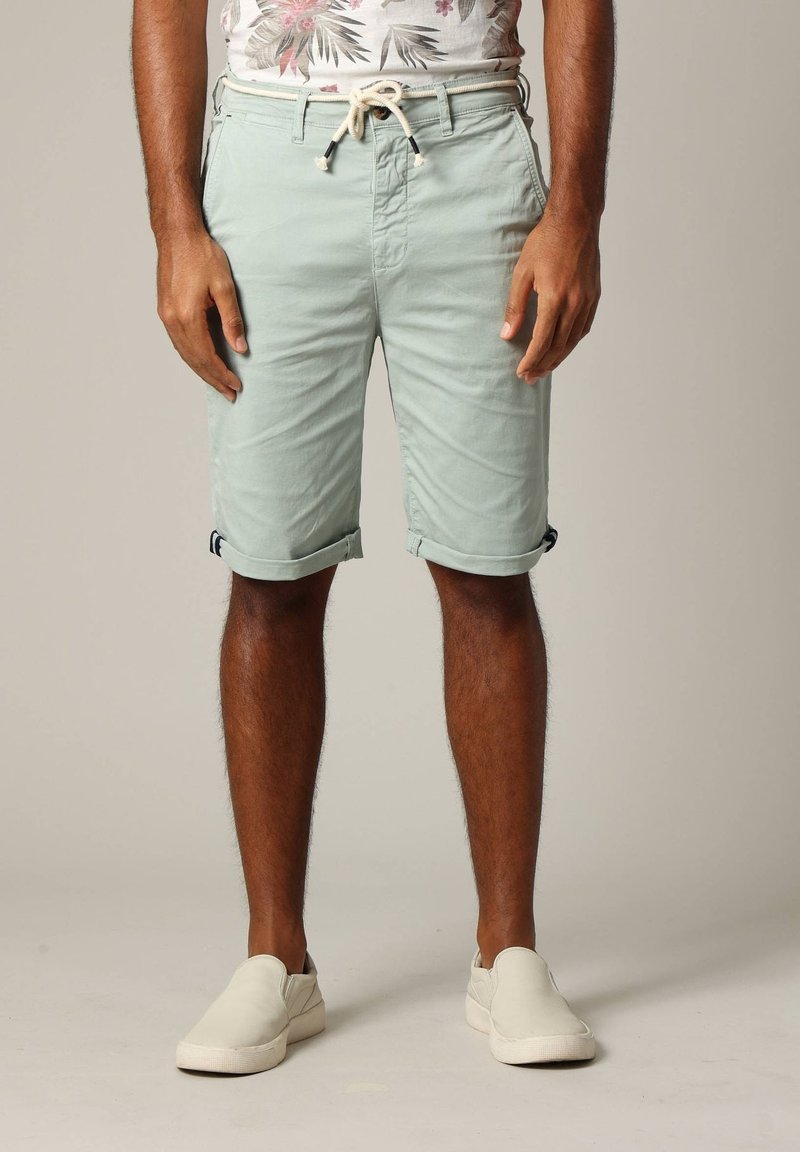 Man in opgerolde lichtgroene shorts met trekkoord en witte instappers, staand tegen een effen achtergrond.