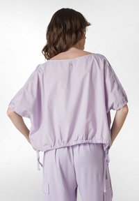 Lila oversized topp med korta ärmar, elastisk fåll och snörningsdetaljer. Slät textur, minimalistisk design och subtil varumärkestagg på baksidan.