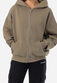 Sudadera con cremallera de color verde oliva, con una gran capucha, bolsillos frontales y puños y dobladillo de canalé. Presenta un discreto logotipo "PEGADOR" en el pecho.