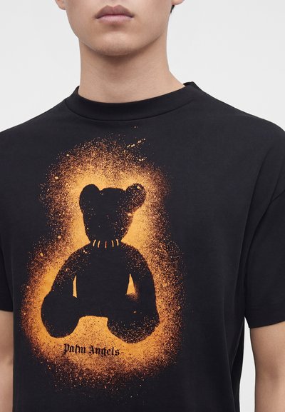 Νέος άνθρωπος φοράει μαύρο t-shirt με γραφικό αρκουδάκι σε στυλ πορτοκαλί σπρέι και το κείμενο "Palm Angels" μπροστά.
