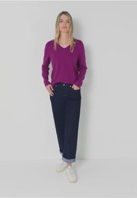 Pull violet à col en V associé à un pantalon bleu marine et des baskets blanches. La tenue présente une coupe décontractée et une texture lisse.
