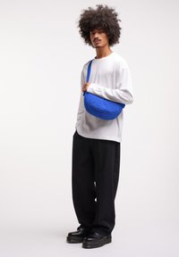 Borsa a tracolla blu con forma arrotondata, realizzata in tessuto morbido. Indossata da una persona in camicia bianca a maniche lunghe e pantaloni neri a gamba larga.
