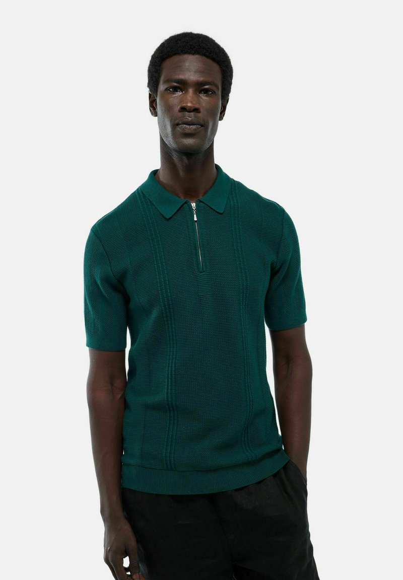 Polo vert à manches courtes avec col zippé, présentant des motifs de tricot texturé et des détails côtelés contrastants autour du col et de l'ourlet.