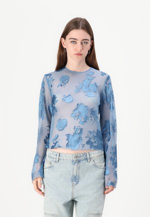 Haut translucide à manches longues bleu clair avec motif floral, présentant un col rond et un design court, associé à un jean taille haute.