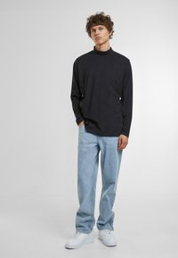 Top lung cu maneci lungi, de culoare închisă, cu guler înalt, asortat cu blugi de culoare albastră deschisă și adidași albi. Textură netedă, croială lejeră. Design simplu.