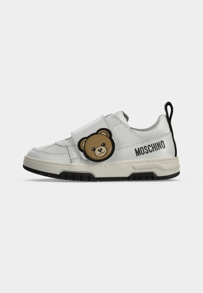 Λευκό αθλητικό παπούτσι Moschino με μαύρη σόλα, λουρί με βέλκρο που έχει καφέ πρόσωπο αρκούδας και μαύρη ταινία έλξης στη φτέρνα.