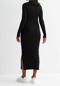 Svart ribbad stickad klänning med lång ärm, en figurnära silhuett och sidofickor; kombinerad med vita sneakers. Slät textur, golvlång.