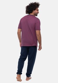 Een man met krullend haar glimlacht, gekleed in een bordeauxrood t-shirt en marineblauwe broek, staande op blote voeten en iets naar de zijkant gedraaid.