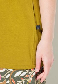Musztardowożółta koszula z krótkim rękawem i bocznym rozcięciem, w towarzystwie wzorzystych zielonych spodni z motywem kwiatowym oraz czarnej metki na koszuli.