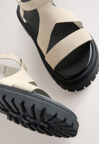 Sandale din piele bej cu talpă neagră, având bretele late încrucișate și o talpă exterioară texturată, robustă, pentru aderență și stabilitate.