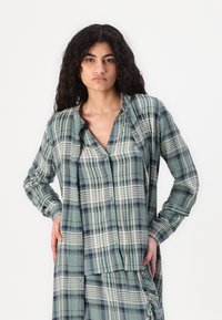 Green plaid button-up shirt with long sleeves, featuring varying stripes and a soft fabric texture. Front pocket and relaxed fit.