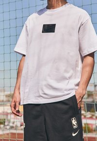 Bílý bavlněný tričko s černým obdélníkovým nášivkou Nike na hrudi, spárované s černými šortkami s logem NBA.
