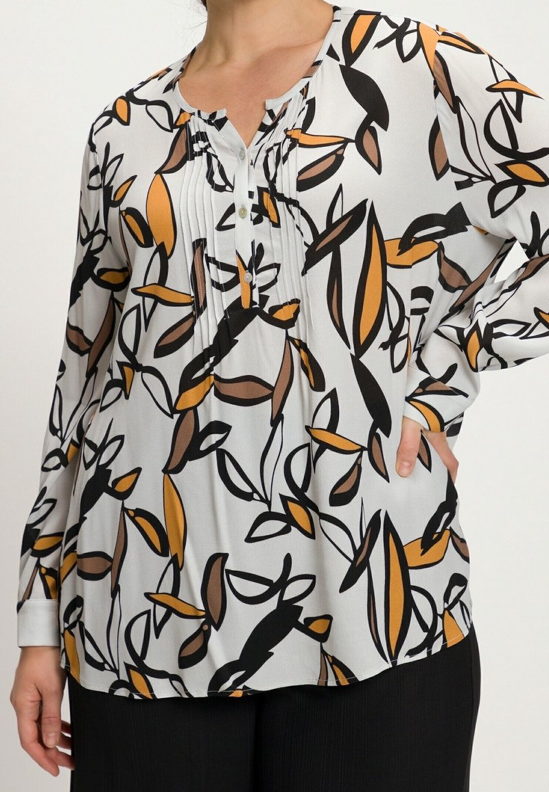 Blouse à manches longues en tissu léger présentant un fond blanc avec des motifs de feuilles noires et orange, une patte de boutonnage et des détails plissés.