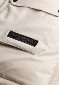 Jasnoróżowy materiał sportowy z teksturowaną czarną metką z napisem "SUPERDRYNYC". Widoczne wzmocnione szwy i zapięcie na zatrzask.