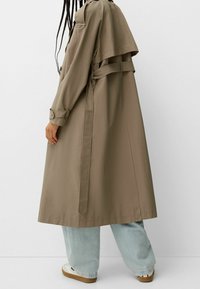 Długi, beżowy płaszcz trencz o dwurzędowym fasonie, z paskiem w talii i naramiennikami, zestawiony z jasnoniebieskimi spodniami i trampkami.