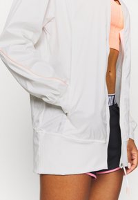 Kevyt valkoinen urheilu takki, jossa on oranssia yksityiskohtia, vetoketjusuljenta ja elastiset hihansuut. Yhdistetty pinkkeihin urheiluShortseihin ja matching yläosaan.