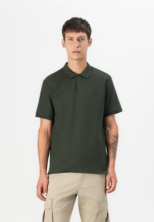 Dark green polo shirt made of textured fabric with a collar, short sleeves, and a subtle logo on the chest. Paired with beige cargo pants.