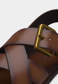 Καφέ δερμάτινο σανδάλι με σταυρωτούς ιμάντες και ορθογώνια χρυσή αγκράφα για ρυθμιζόμενο κούμπωμα.