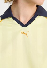 Gul sportspolo med navy krage, försedd med vertikala ränder och en guld Puma-logotyp på bröstet. Materialet verkar texturerat och andas.