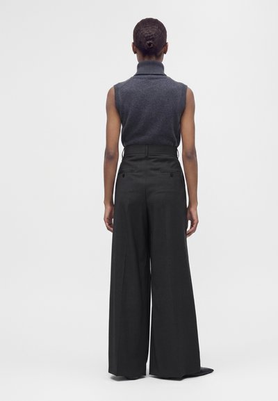 Personne debout dos à la caméra, portant un col roulé sans manches gris foncé et un pantalon noir taille haute à jambes larges.
