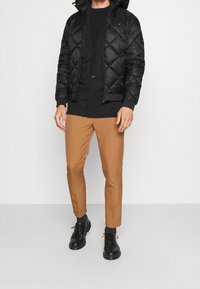 Svart, vadderad vinterjacka med huva, lagd över en svart tröja och bruna byxor. Skorna är svarta med snören och texturerade strumpor.