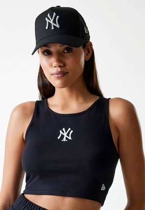 Femme portant une casquette noire des New York Yankees et un crop top assorti avec le logo de l'équipe sur un fond blanc uni.