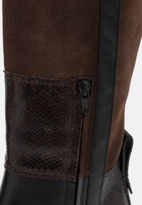 Gros plan sur une botte en suède marron et cuir noir avec un patch en texture de peau de serpent et une fermeture éclair sur le côté.