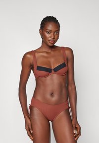 Bikini komplet v rjavi barvi z črnimi detajli. Zgornji del ima široke naramnice in žične košarice, medtem ko ima spodnji del nastavljive strani in rebrasto teksturo.