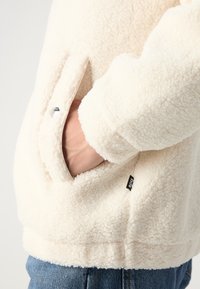 Creme fleece-jakke med en tekstureret overflade, der har en sidelomme og et logo-tag. Ærmerne har en afslappet pasform og er let oversize.
