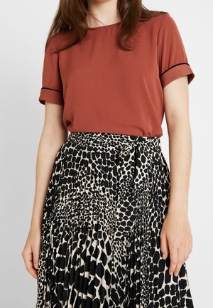 Femme portant un haut à manches courtes couleur rouille, rentré dans une jupe plissée à imprimé animal noir et blanc avec une ceinture à la taille.