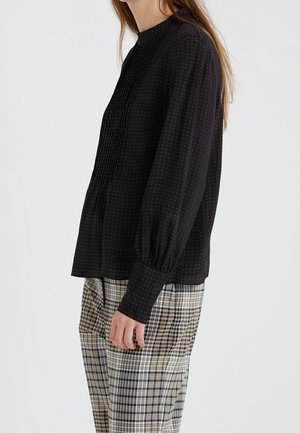 Schwarze Bluse mit langen Ärmeln, Falten und hohem Kragen. Aus strukturiertem Stoff mit kleinen bunten Punkten. Kombiniert mit karierter Hose.