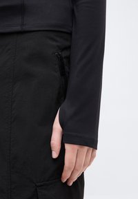 Χέρι που ακουμπάει δίπλα σε μαύρο παντελόνι με φερμουάρ στην τσέπη, φορώντας μαύρο μακρυμάνικο τοπ με άνοιγμα για τον αντίχειρα, σε ανοιχτό φόντο.