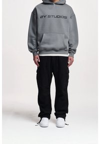 Sudadera con capucha gris, bolsillo frontal y texto negro "2Y STUDIOS"; combinada con pantalones cargo negros y zapatillas blancas. Estilo sencillo y ajuste relajado.