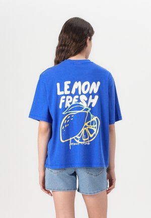 Person iført en blå t-shirt med teksten "LEMON FRESH" og citrongrafik på ryggen, kombineret med lyse denimshorts.