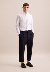 Witte knoopshirt met lichtblauwe strepen, gecombineerd met donkerblauwe geplooide broek en zwarte leren schoenen. Eenvoudig zakdetail op het shirt.