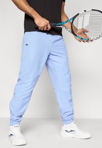 Αθλητικό παντελόνι ανοιχτού γαλάζιου χρώματος με ελαστικά μανσέτα, διακοσμημένο με πράσινο κεντητό λογότυπο. Φορεμένο με λευκά αθλητικά παπούτσια και κρατώντας ρακέτα τένις.