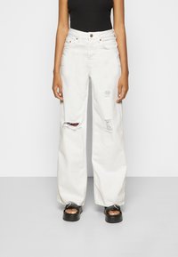 Calças largas brancas feitas de denim com detalhes desgastados. Corte de cintura alta, com fecho de botão e bolsos. Combinadas com sapatos de plataforma pretos.