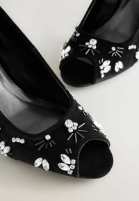 Cizme cu toc deschis negre din catifea, ornamentate cu strălucitoare cristale și accente negre, având o siluetă elegantă și o textură netedă.