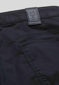 Pantalon en mélange de coton noir avec une étiquette en cuir rectangulaire portant l'inscription "M5", avec des coutures visibles et des poches arrière. Texture lisse.