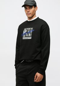 Felpa nera con maniche lunghe, caratterizzata da una stampa grafica e un testo in blu e bianco. Indossata con pantaloni scuri e un cappellino.