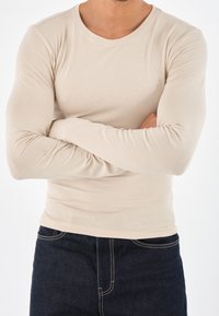 Beige långärmad skjorta i mjukt tyg, figursydd design, rund halsringning, med armarna i kors mot en enfärgad vit bakgrund.